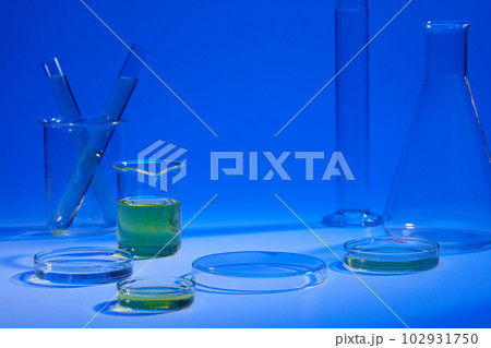 Beaker and petri dish of yellow liquid displayed with other laboratory glassware on a blue background. Vacant space on podium Beaker and petri dish of yellow liquid displayed with other laboratory glassware on a blue background. Vacant space on podium 102931750