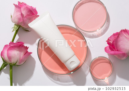 Scene for branding cosmetic of rose extract, plastic tube unlabeled for design placed on petri dish containing pink liquid and fresh roses arranged on white background 102932515