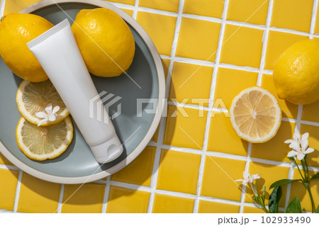 Empty label white tube, Lemon slices and small flower put on a round dish. Lemon (Citrus limon) can reduce inflammation and excess sebum production. Yellow mosaic tiles background 102933490