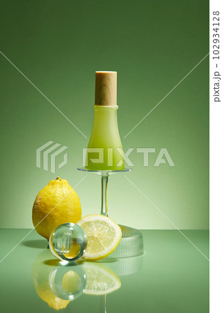 Green background for branding and minimal presentation of product from lemon ingredient. Glass bottle unlabeled on glass cup upside down decorated with glass ball and fresh lemon. Mockup, copy space Green background for branding and minimal presentation of product from lemon ingredient. Glass bottle unlabeled on glass cup upside down decorated with glass ball and fresh lemon. Mockup, copy space 102934128