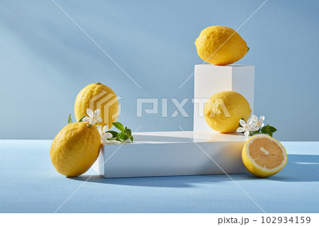 Pedestal for display cosmetic product of lemon extract with white empty podiums, lemons and white flowers on blue background. Lemon has many good uses for health and beauty. Pedestal for display cosmetic product of lemon extract with white empty podiums, lemons and white flowers on blue background. Lemon has many good uses for health and beauty. 102934159