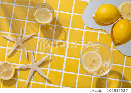 Lemon placed on a mirror arranged with starfishes and two glasses containing Lemon slices. Vacant space on the yellow mosaic tiles background to show natural cosmetic product 102935527