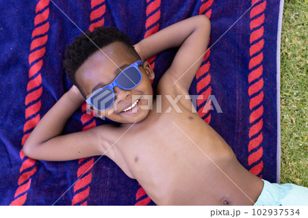 High angle view of african american smiling shirtless boy wearing sunglasses and lying on towel High angle view of african american smiling shirtless boy wearing sunglasses and lying on towel 102937534
