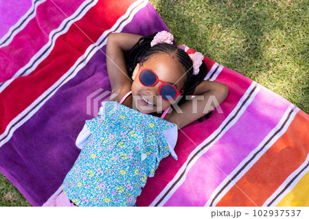Cute african american girl wearing sunglasses with hands behind head lying on towel 102937537