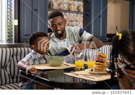 African american father pouring milk in bowl for son while having breakfast at dining table 102937641