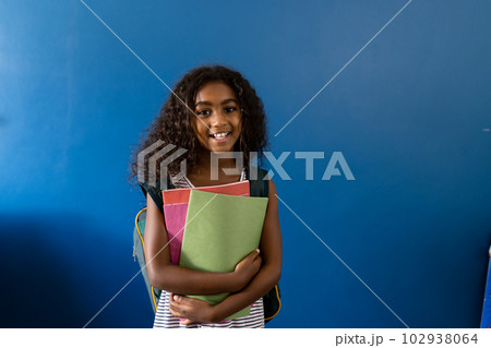 Portrait of happy biracial schoolgirl with school bag holding notebooks on blue background 102938064