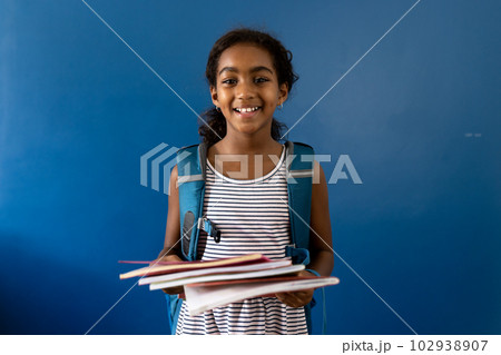 Portrait of happy biracial schoolgirl holding notebooks smiling, on blue background with copy space Portrait of happy biracial schoolgirl holding notebooks smiling, on blue background with copy space 102938907
