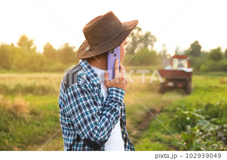 Farmer talking on the purple phone on the tractor background. Amidst the vast expanse of a field, a farmer confidently walks, seamlessly multitasking by engaging in a conversation on mobile phone Farmer talking on the purple phone on the tractor background. Amidst the vast expanse of a field, a farmer confidently walks, seamlessly multitasking by engaging in a conversation on mobile phone 102939049