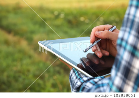 Tablet screen and stylus. An agronomist farmer man is seen using a digital tablet computer amidst a young cornfield during the serene hours of sunrise or sunset. Close up hands Tablet screen and stylus. An agronomist farmer man is seen using a digital tablet computer amidst a young cornfield during the serene hours of sunrise or sunset. Close up hands 102939051