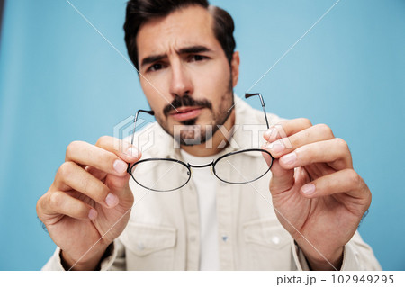 Close-up portrait of a man looking through glasses that he holds in his hands, dioptric lenses, glasses for far-sightedness and near-sightedness, on a blue background in a white T-shirt, copy space  102949295