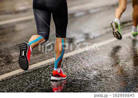 legs female runner athlete run marathon race on wet asphalt, blue kinesio tape on shins legs female runner athlete run marathon race on wet asphalt, blue kinesio tape on shins 102950845