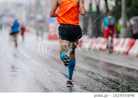rear view runner athlete run marathon race on rain, in blue compression sleeve on feet and orange singlet 102950846