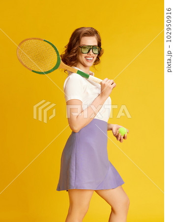 Portrait of smiling, young, beautiful girl in sportswear and glasses, posing with tennis racket and ball against yellow studio background Portrait of smiling, young, beautiful girl in sportswear and glasses, posing with tennis racket and ball against yellow studio background 102950916