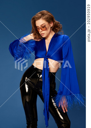 Portrait of stylish, beautiful young woman in trendy sunglasses, top and leather pants posing against blue studio background Portrait of stylish, beautiful young woman in trendy sunglasses, top and leather pants posing against blue studio background 102950919