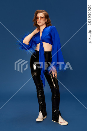 Portrait of stylish, beautiful young woman in trendy sunglasses, top and leather pants posing against blue studio background Portrait of stylish, beautiful young woman in trendy sunglasses, top and leather pants posing against blue studio background 102950920