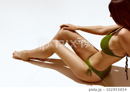 Cropped image of tanned female body with spf cream on legs. Model with slim, tanned skin posing in swimsuit against white background 102953854