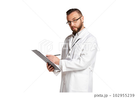 Portrait of concentrated young man, doctor in white lab coat standing, putting information in medical papers against white background Portrait of concentrated young man, doctor in white lab coat standing, putting information in medical papers against white background 102955239
