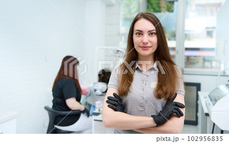 Confident portrait of a young female doctor dentist hygienist, healthcare worker, dental assistant posing with her arms folded, looking at camera, standing in her workplace in modern dentistry clinic 102956835