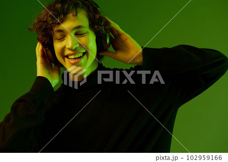 Man teenager wearing headphones listening to music, dancing and singing with his eyes closed, DJ party happiness and smile, hipster lifestyle, portrait green background mixed neon light, copy space Man teenager wearing headphones listening to music, dancing and singing with his eyes closed, DJ party happiness and smile, hipster lifestyle, portrait green background mixed neon light, copy space 102959166