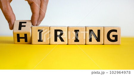 Hiring or firing symbol. Businessman hand turns a wooden cube and changes the word 'firing' to 'hiring'. Beautiful yellow table, white background, copy space. Business and hiring or firing concept. Hiring or firing symbol. Businessman hand turns a wooden cube and changes the word 'firing' to 'hiring'. Beautiful yellow table, white background, copy space. Business and hiring or firing concept. 102964978