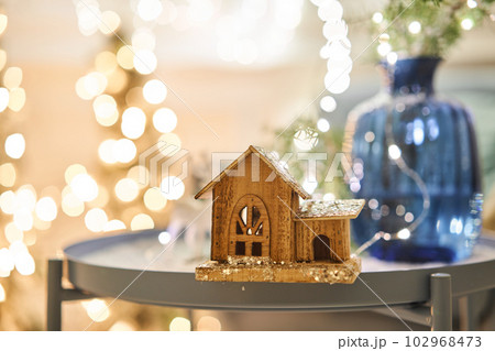 Decorated table in the Christmas interior. Christmas tree. Close-up a small house, Cones, garland, and other Christmas tree toys. light from the garlands Decorated table in the Christmas interior. Christmas tree. Close-up a small house, Cones, garland, and other Christmas tree toys. light from the garlands 102968473