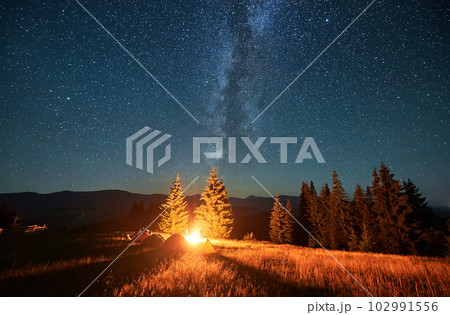 Night camping in mountains under starry sky. Tourist tents in campsite under beautiful sky full of stars with Milky way. Silhouette of hikers having a rest near burning campfire. Concept of tourism. Night camping in mountains under starry sky. Tourist tents in campsite under beautiful sky full of stars with Milky way. Silhouette of hikers having a rest near burning campfire. Concept of tourism. 102991556