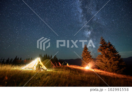 Night camping in mountains under starry sky. Male tourist sitting near tent in campsite, having rest, admiring landscape and fire. Man enjoying beautiful night sky full of stars with Milky way. Night camping in mountains under starry sky. Male tourist sitting near tent in campsite, having rest, admiring landscape and fire. Man enjoying beautiful night sky full of stars with Milky way. 102991558