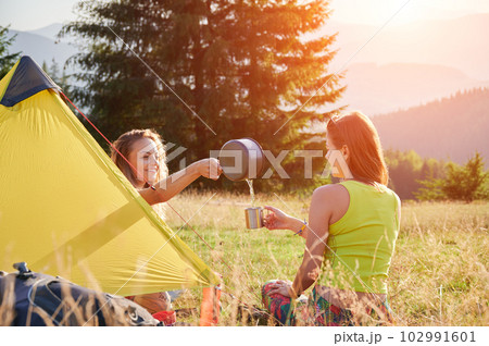 Two women travellers camping outdoors. Young women sitting in campsite, having break. Friends sitting on grass, talking, smiling, pouring water, making tea. Concept of tourism and hiking in mountains. 102991601