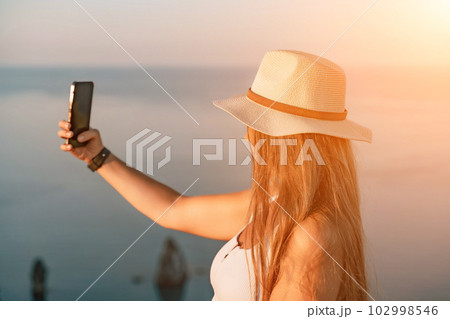 Selfie woman in a hat, white tank top, and shorts captures a selfie shot with her mobile phone against the backdrop of a serene beach and blue sea. 102998546