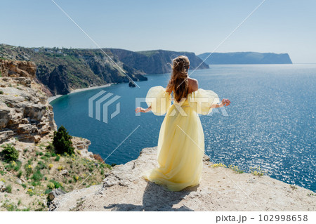 Woman in a yellow dress on the sea. Side view Young beautiful sensual woman in yellow long dress posing on a rock high above the sea at sunset. Girl in nature against the blue sky 102998658