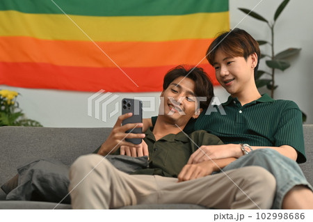 Beautiful moment of happy same sex male couple checking social media on smartphone while relaxing on couch at home 102998686