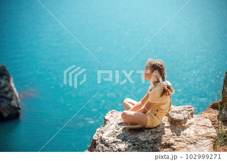 Happy girl perched atop a high rock above the sea, wearing a yellow jumpsuit and braided hair, signifying the concept of summer vacation at the beach. Happy girl perched atop a high rock above the sea, wearing a yellow jumpsuit and braided hair, signifying the concept of summer vacation at the beach. 102999271