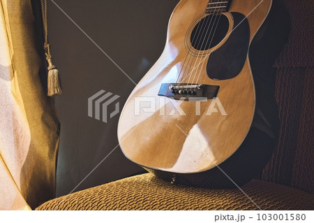 A Spanish acoustic classical guitar with steel and nylon strings resting on a chair next to a window curtain 103001580
