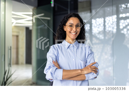 Portrait of happy and successful business woman, boss in shirt smiling and looking at camera inside office with crossed arms, Hispanic woman with curly hair in corridor. 103005336
