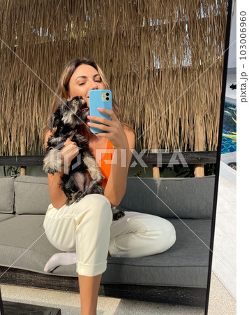 Woman in orange top and cosy comfy pants at home outdoor take photo selfie on phone with puppy miniature schnauzer in mirror for social media, stories, vertical Woman in orange top and cosy comfy pants at home outdoor take photo selfie on phone with puppy miniature schnauzer in mirror for social media, stories, vertical 103006960