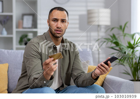 Worried young african american man sitting on couch at home and looking upset at camera, holding credit card and phone. Problems with the account, transaction, online purchase. 103010939