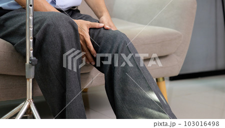 Senior grandfather with walking stick trying to exercise knee and leg on sofa 103016498
