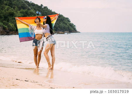 Couple of cheerful women with rainbow flag standing on beach 103017019