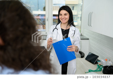 Confident portrait of a Caucasian charming pretty woman, female doctor, GP, physician, gynecologist in white medical gown, smiling broadly, welcoming her patient at appointment in new medical clinic 103034837
