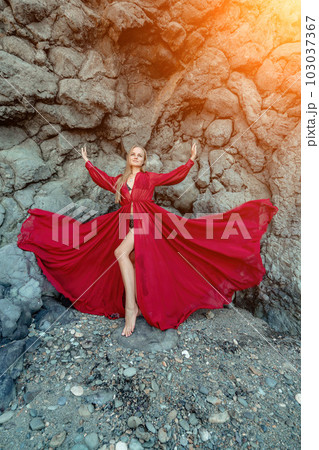 Red dress rocks woman. A blonde with flowing hair in a long flowing red dress stands near a rock of volcanic origin. Travel concept, photo session at sea Red dress rocks woman. A blonde with flowing hair in a long flowing red dress stands near a rock of volcanic origin. Travel concept, photo session at sea 103037367