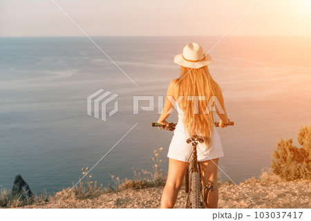 Woman travel bike sea. Happy woman cyclist sitting on her bike, enjoying the beautiful mountain and sea landscape, signifying the idea of an adventurous bike ride. 103037417