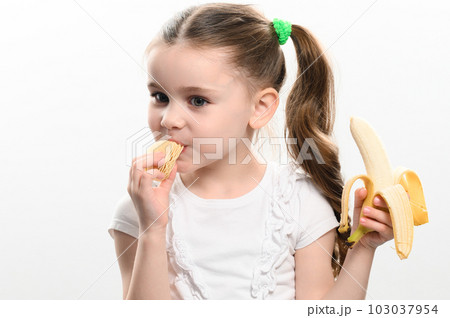 A little girl is eating potato chips and holding a banana 103037954
