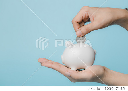 Hand saving a coin into piggy bank on blue background Hand saving a coin into piggy bank on blue background 103047136