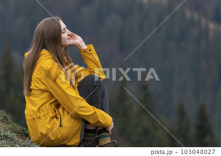 Pensive woman looking into the distance. Young pretty girl wears yellow raincoat on forest background. 103048027