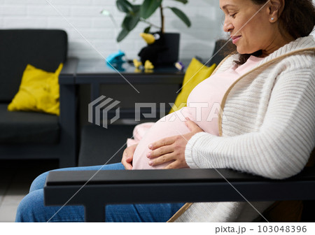 Close-up cheerful adult pregnant woman smiling, touching her belly, enjoying her pregnancy, feeling baby movements in womb, sitting relaxed on sofa in modern interior. Happy carefree pregnancy concept 103048396