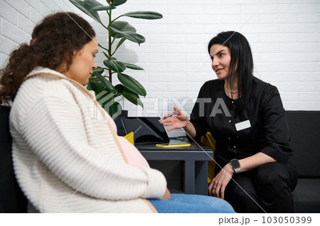 Adult pregnant female patient talking with doctor about test result diagnosis and ultrasound scan of baby at hospital corridor, listening to doctor's prescription. Obstetrics and gynecology concept Adult pregnant female patient talking with doctor about test result diagnosis and ultrasound scan of baby at hospital corridor, listening to doctor's prescription. Obstetrics and gynecology concept 103050399