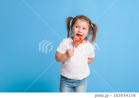 Beautiful little female child holding huge lollipop spiral candy smiling happy isolated on blue background. Beautiful little female child holding huge lollipop spiral candy smiling happy isolated on blue background. 103054139