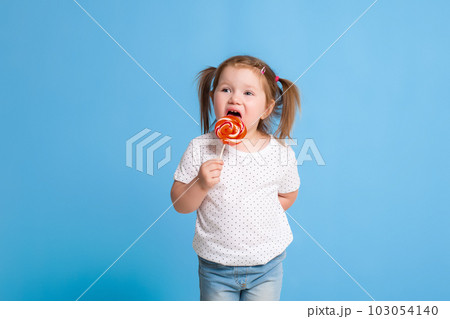 Beautiful little female child holding huge lollipop spiral candy smiling happy isolated on blue background. Beautiful little female child holding huge lollipop spiral candy smiling happy isolated on blue background. 103054140