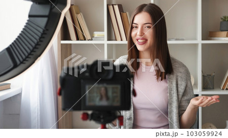 Video vlogging. Online coaching. Attractive happy woman blogger creating internet content talking recording interview on camera on tripod ring light home interior. 103059924