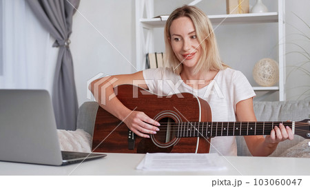 Online guitar class. Music tutor. Smiling pleased woman teacher tutorial lesson teaching playing instrument recording educational video on laptop camera home interior. 103060047